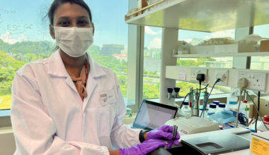Photo of Shruthi Pandi Chelvam in lab, using a piece of lab equipment in front of a large window with treetops and some buildings in the background.